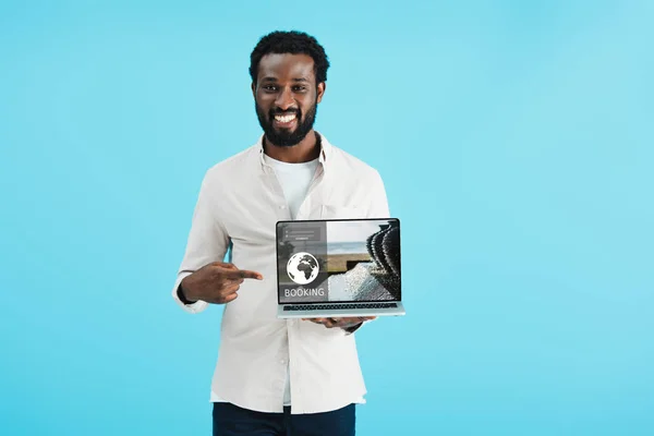 Lächelnder afrikanisch-amerikanischer Mann zeigt auf Laptop mit Buchungswebseite isoliert auf blau — Stockfoto