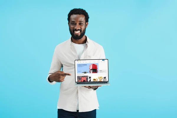 KYIV, UKRAINE - 17 MAI 2019 : homme afro-américain souriant pointant vers un ordinateur portable avec le site Web ebay, isolé sur bleu — Photo de stock