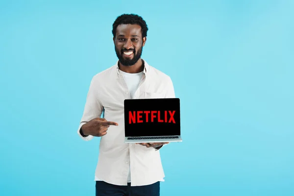 KYIV, UCRANIA - 17 de mayo de 2019: sonriente hombre afroamericano apuntando a la computadora portátil con el sitio web de netflix, aislado en azul — Stock Photo