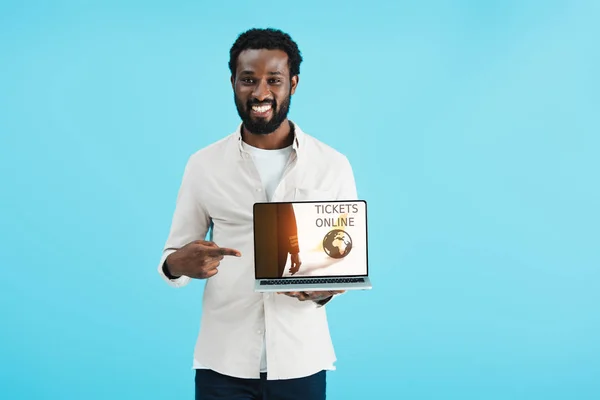 Lächelnder afrikanisch-amerikanischer Mann zeigt auf Laptop mit Tickets Online-Website isoliert auf blau — Stockfoto