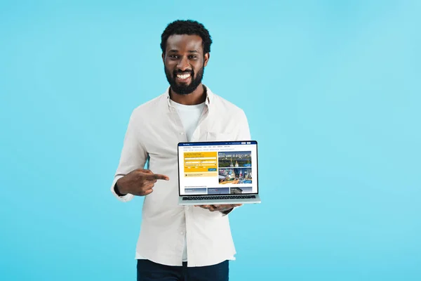 Kyiv, ukraine - 17. Mai 2019: lächelnder afrikanisch-amerikanischer Mann zeigt auf Laptop mit Buchungswebseite, isoliert auf blau — Stockfoto