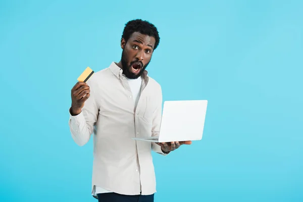 Choqué homme afro-américain shopping en ligne avec carte de crédit et ordinateur portable isolé sur bleu — Photo de stock