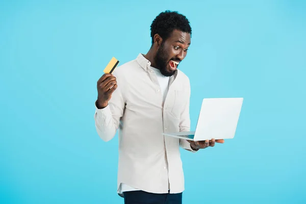 Збуджений афроамериканський чоловік купує онлайн з кредитною карткою та ноутбуком ізольовано на синьому — стокове фото