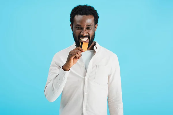 Smiling african american man biting credit card isolated on blue — Stock Photo