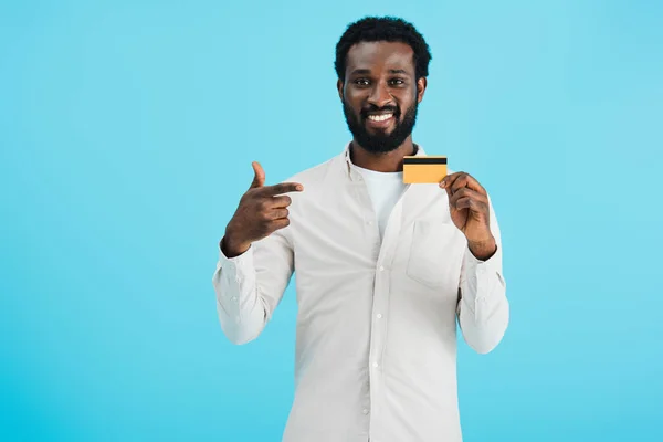 Guapo sonriente afroamericano hombre apuntando a la tarjeta de crédito aislado en azul - foto de stock