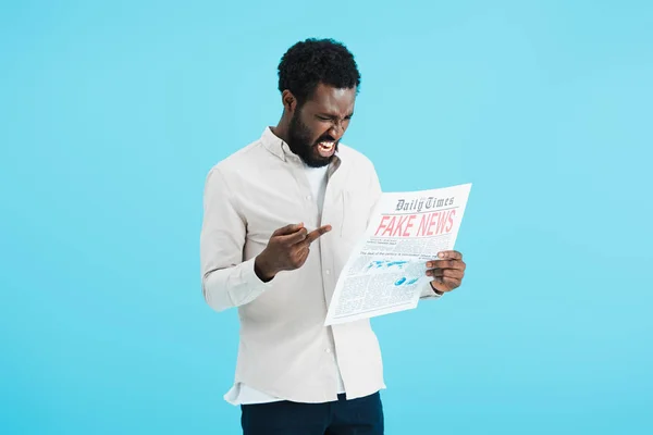 Aggressive african american man reading newspaper with fake news and showing middle finger isolated on blue — Stock Photo