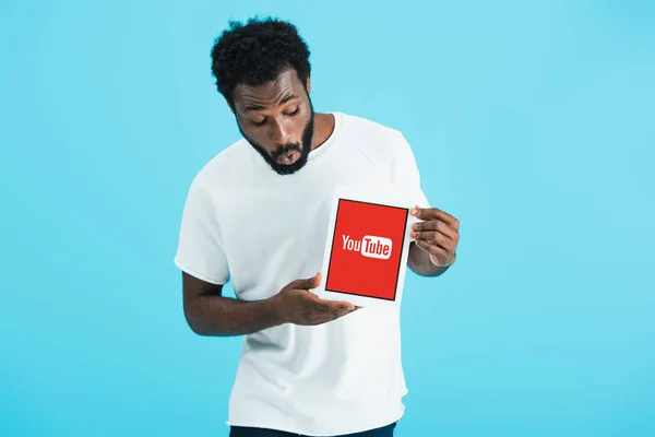 KYIV, UKRAINE - 17 MAI 2019 : un homme afro-américain choqué regardant une tablette numérique avec une application youtube, isolé sur bleu — Photo de stock