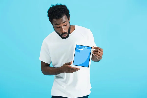 KYIV, UCRANIA - 17 de mayo de 2019: sorprendido hombre afroamericano mirando la tableta digital con la aplicación de twitter, aislado en azul - foto de stock