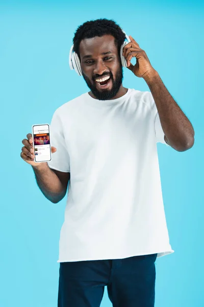 KYIV, UCRAINA - 17 MAGGIO 2019: uomo afroamericano sorridente che ascolta musica con le cuffie e mostra smartphone con app soundcloud, isolato sul blu — Foto stock