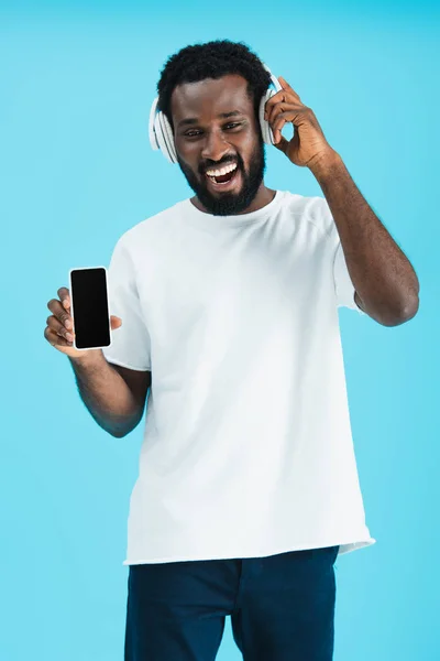 Allegro uomo afroamericano che ascolta musica con cuffie e mostra smartphone con schermo bianco, isolato su blu — Foto stock