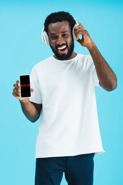 KYIV, UCRAINA - 17 MAGGIO 2019: uomo afroamericano sorridente che ascolta musica con le cuffie e mostra smartphone con app netflix, isolato sul blu — Foto stock