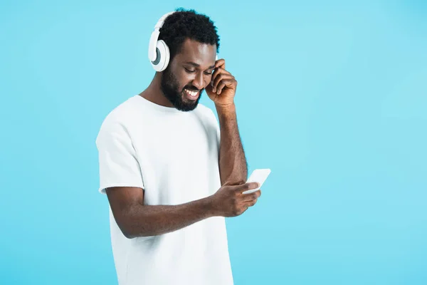 Homem americano africano alegre ouvindo música com fones de ouvido e usando smartphone, isolado em azul — Fotografia de Stock