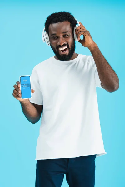KYIV, UCRAINA - 17 MAGGIO 2019: uomo afroamericano sorridente che ascolta musica con le cuffie e mostra smartphone con app skype, isolato su blu — Foto stock