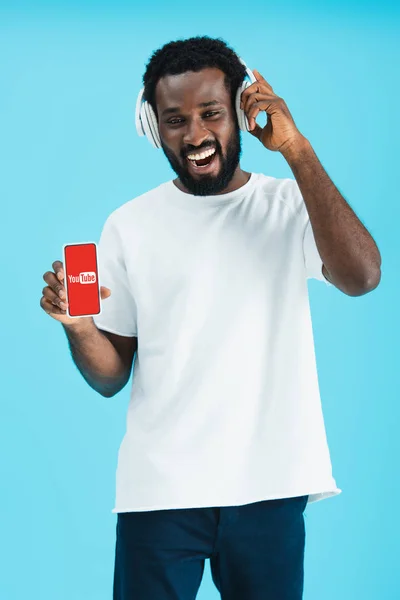 KYIV, UKRAINE - 17 MAI 2019 : homme afro-américain souriant écoutant de la musique avec écouteurs et montrant smartphone avec application youtube, isolé sur bleu — Photo de stock