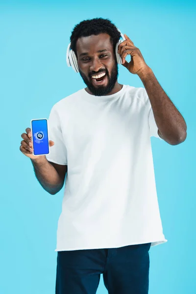 KYIV, UCRAINA - 17 MAGGIO 2019: uomo afroamericano sorridente che ascolta musica con le cuffie e mostra smartphone con app shazam, isolato sul blu — Foto stock