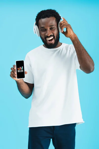 КИЕВ, УКРАИНА - 17 мая 2019 года: улыбающийся африканский американец слушает музыку в наушниках и показывает смартфон с приложением Deezer, изолированным на синем — стоковое фото