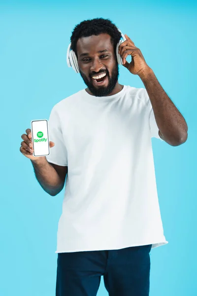 KYIV, UCRANIA - 17 de mayo de 2019: hombre afroamericano sonriente escuchando música con auriculares y mostrando teléfono inteligente con aplicación spotify, aislado en azul - foto de stock