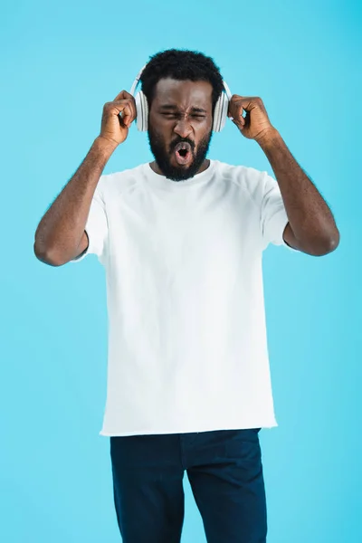 Animado afro-americano homem cantando e ouvindo música com fones de ouvido, isolado em azul — Fotografia de Stock