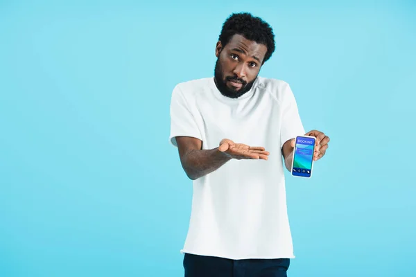 Frustrado hombre afroamericano mostrando teléfono inteligente con aplicación de reserva, aislado en azul - foto de stock
