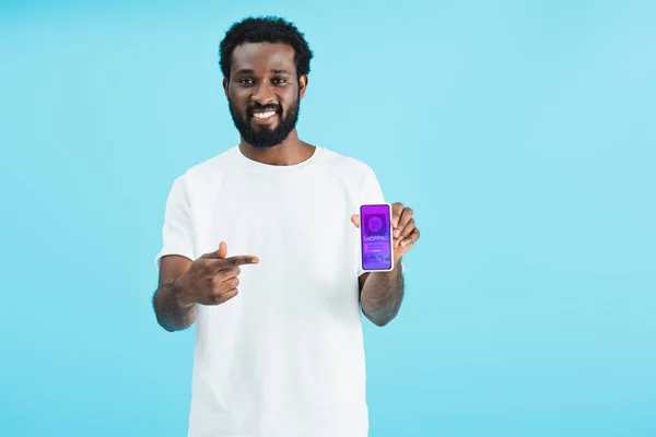 Uomo afroamericano sorridente che punta verso smartphone con app per lo shopping, isolato sul blu — Foto stock