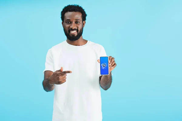 КИЕВ, УКРАИНА - 17 мая 2019 года: улыбающийся африканский американец указывает на смартфон с помощью приложения shazam, изолированного на синем — стоковое фото