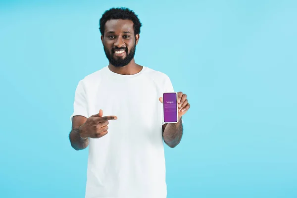 KYIV, UKRAINE - MAY 17, 2019: smiling african american man pointing at smartphone with instagram app, isolated on blue — Stock Photo