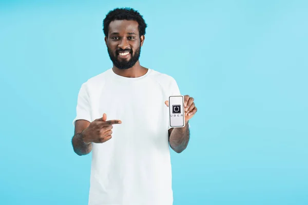 KYIV, UKRAINE - MAY 17, 2019: smiling african american man pointing at smartphone with uber app, isolated on blue — Stock Photo