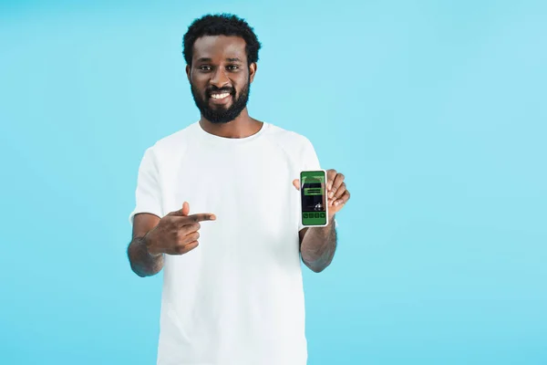 Lächelnder afrikanischer Amerikaner zeigt auf Smartphone mit Buchungs-App, isoliert auf blauem Grund — Stockfoto