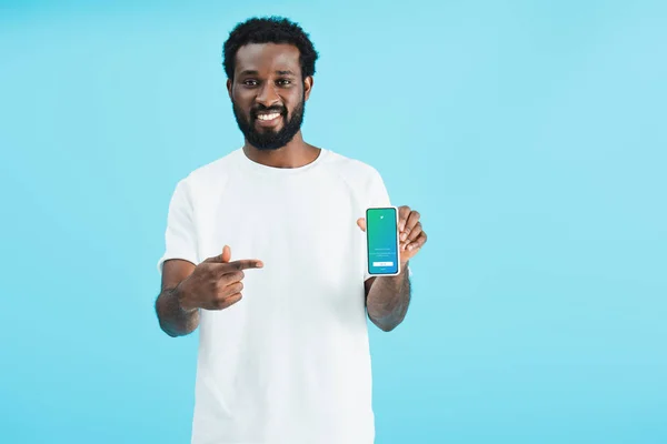 КИЕВ, УКРАИНА - 17 мая 2019 года: улыбающийся африканский американец указывает на смартфон с приложением twitter, изолированным на синем — стоковое фото