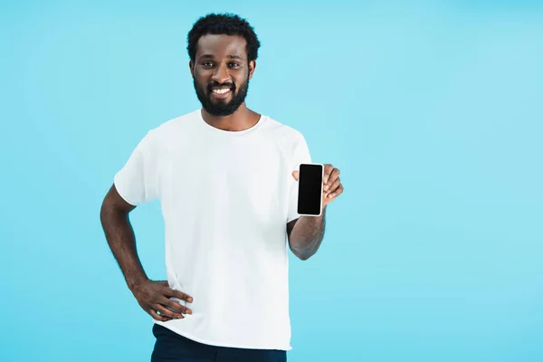 Sonriente hombre afroamericano apuntando a teléfono inteligente con pantalla en blanco, aislado en azul - foto de stock