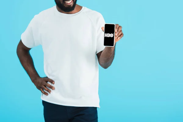 KYIV, UCRANIA - 17 de mayo de 2019: vista recortada del hombre afroamericano mostrando su teléfono inteligente con la aplicación HBO, aislado en azul - foto de stock
