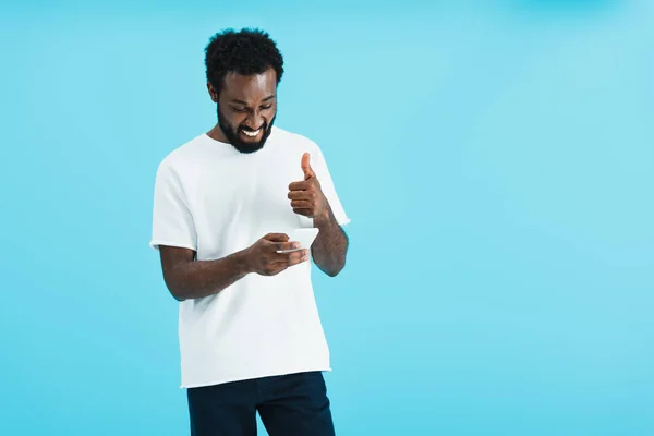 Hombre afroamericano sonriente con signo de ok usando teléfono inteligente, aislado en azul - foto de stock