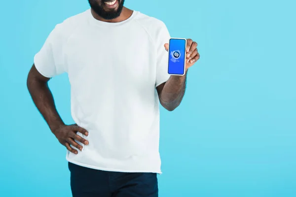 KYIV, UCRANIA - 17 de mayo de 2019: vista recortada del hombre afroamericano que muestra el teléfono inteligente con la aplicación shazam, aislado en azul - foto de stock