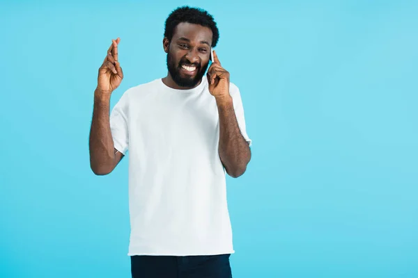 Sonriente hombre afroamericano con los dedos cruzados hablando en el teléfono inteligente, aislado en azul - foto de stock