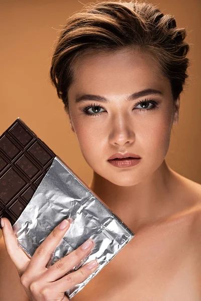 Hermosa mujer desnuda joven sosteniendo barra de chocolate en lámina de plata aislada en beige - foto de stock