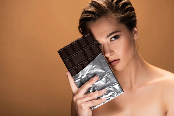 Bella giovane donna nuda con barretta di cioccolato in lamina d'argento vicino al viso isolato sul beige — Foto stock