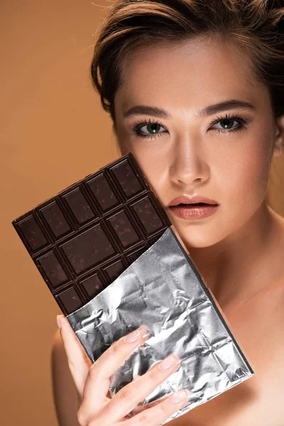 Молода гола жінка тримає шоколадний батончик у срібній фользі біля обличчя ізольовано на бежевому — стокове фото