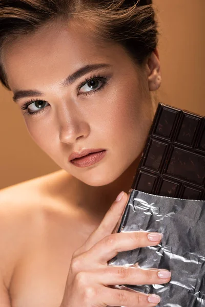 Beautiful young nude woman holding chocolate bar in silver foil isolated on beige — Stock Photo