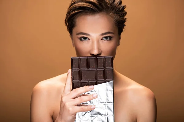 Belle jeune femme nue tenant barre de chocolat en feuille d'argent devant la bouche isolée sur beige — Photo de stock