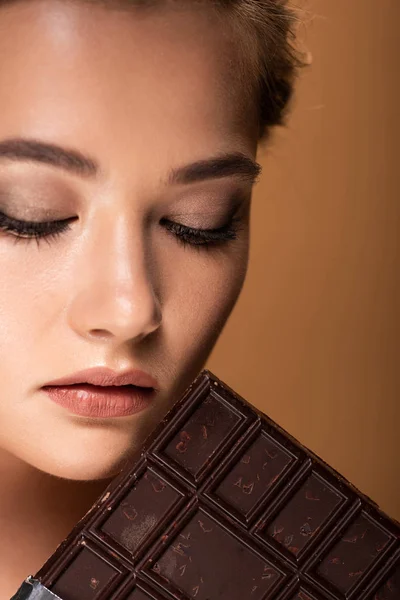 Hermosa joven con los ojos cerrados sosteniendo barra de chocolate aislado en beige - foto de stock