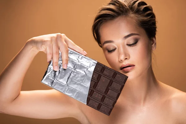 Красива молода гола жінка з закритими очима тримає шоколадний батончик у срібній фользі ізольована на бежевому — стокове фото