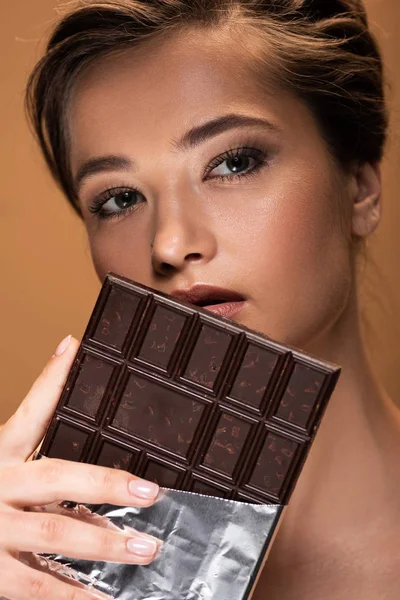 Bela jovem segurando barra de chocolate em folha de prata perto do rosto isolado no bege — Fotografia de Stock