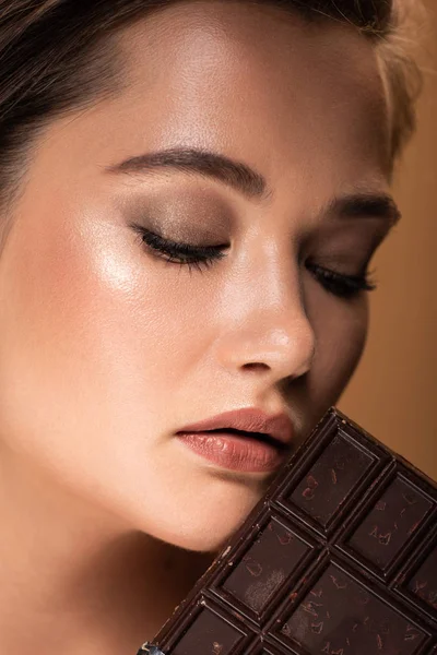 Bella giovane donna con gli occhi chiusi che tiene la barra di cioccolato vicino al viso isolato sul beige — Foto stock