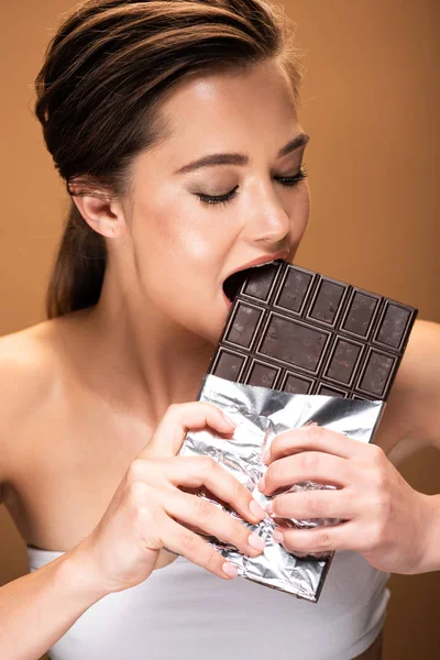 Молодая женщина кусает шоколад в серебряной фольге изолированы на бежевый — стоковое фото