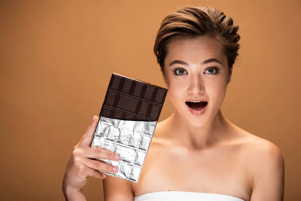 Beautiful excited young woman holding chocolate bar in silver foil isolated on beige — Stock Photo