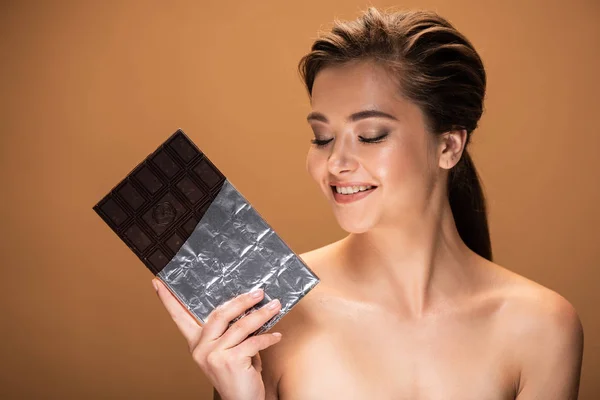 Beautiful smiling young naked woman holding chocolate bar in silver foil isolated on beige — Stock Photo