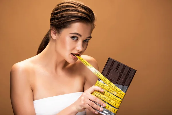 Beautiful young woman holding measuring tape in mouth and chocolate bar in silver foil while looking at camera isolated on beige — Stock Photo