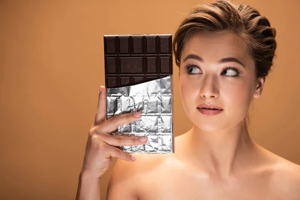 Beautiful young nude woman looking at chocolate bar in silver foil isolated on beige — Stock Photo
