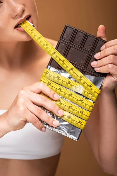 Partial view of woman with yellow measuring tape in mouth holding chocolate bar isolated on beige — Stock Photo