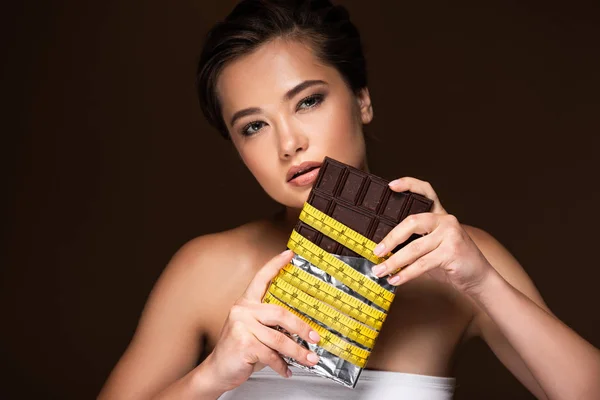 Young woman holding chocolate bar in yellow measuring tape isolated on black — Stock Photo